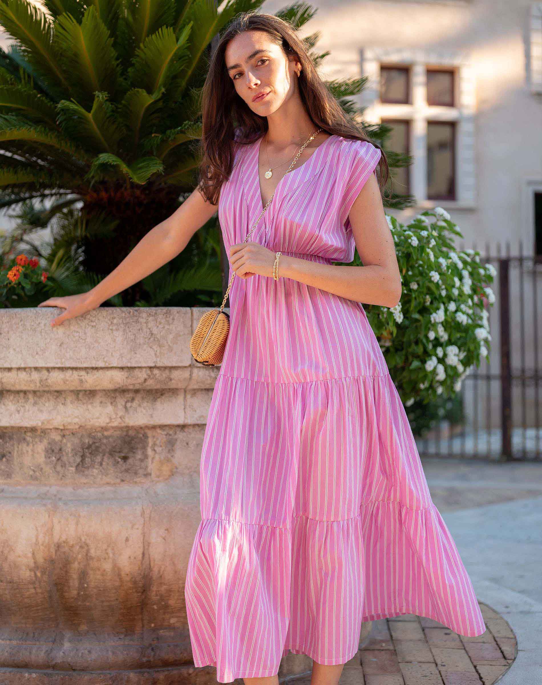Woman in a pink dress standing outdoors with plants and a building in the background