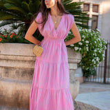 Woman in a pink dress standing outdoors with plants and a building in the background