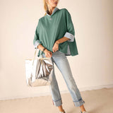 Woman wearing a green shirt, light blue jeans, and white sneakers, holding a silver bag against a beige background.
