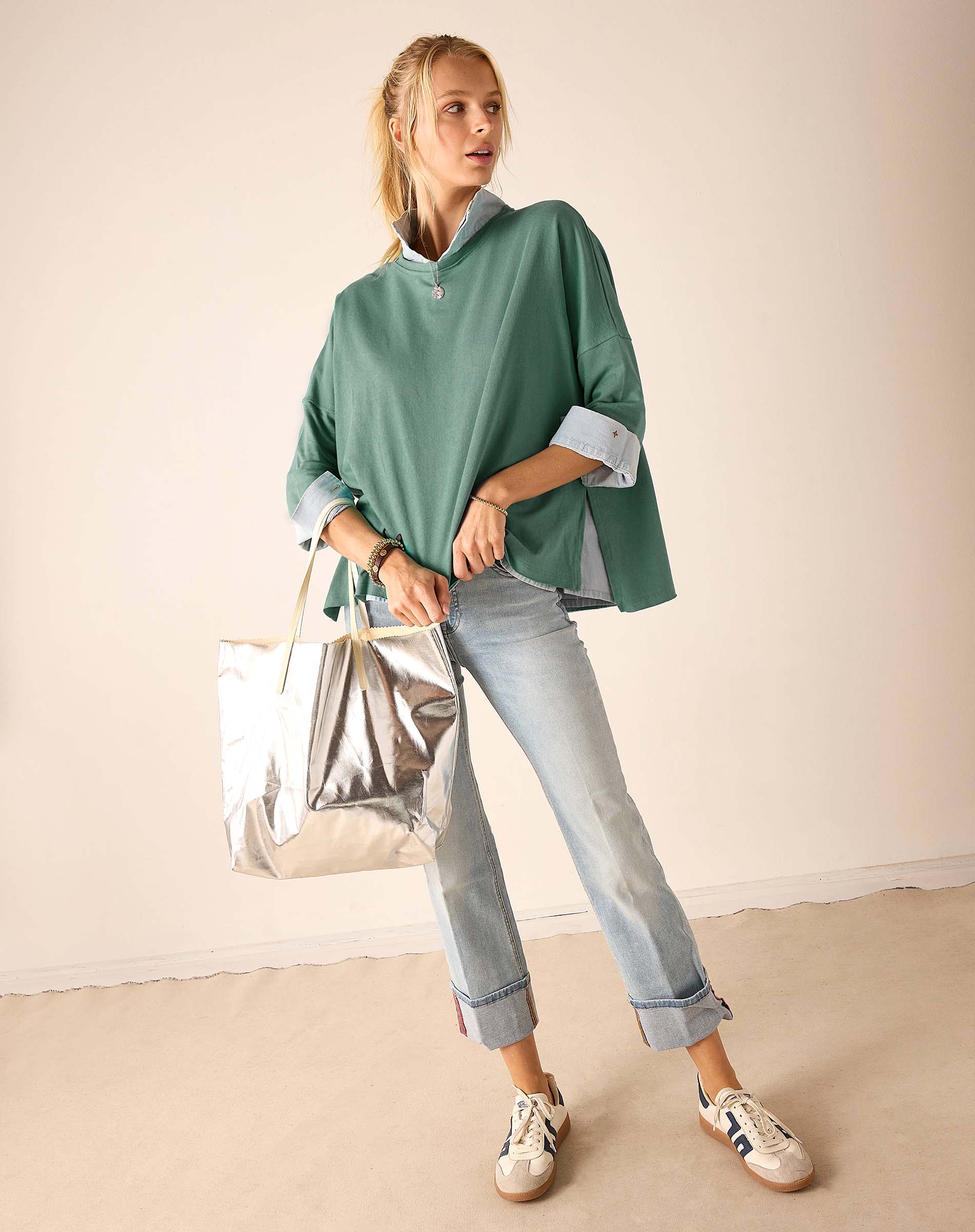 Woman wearing a green shirt, light blue jeans, and white sneakers, holding a silver bag against a beige background.