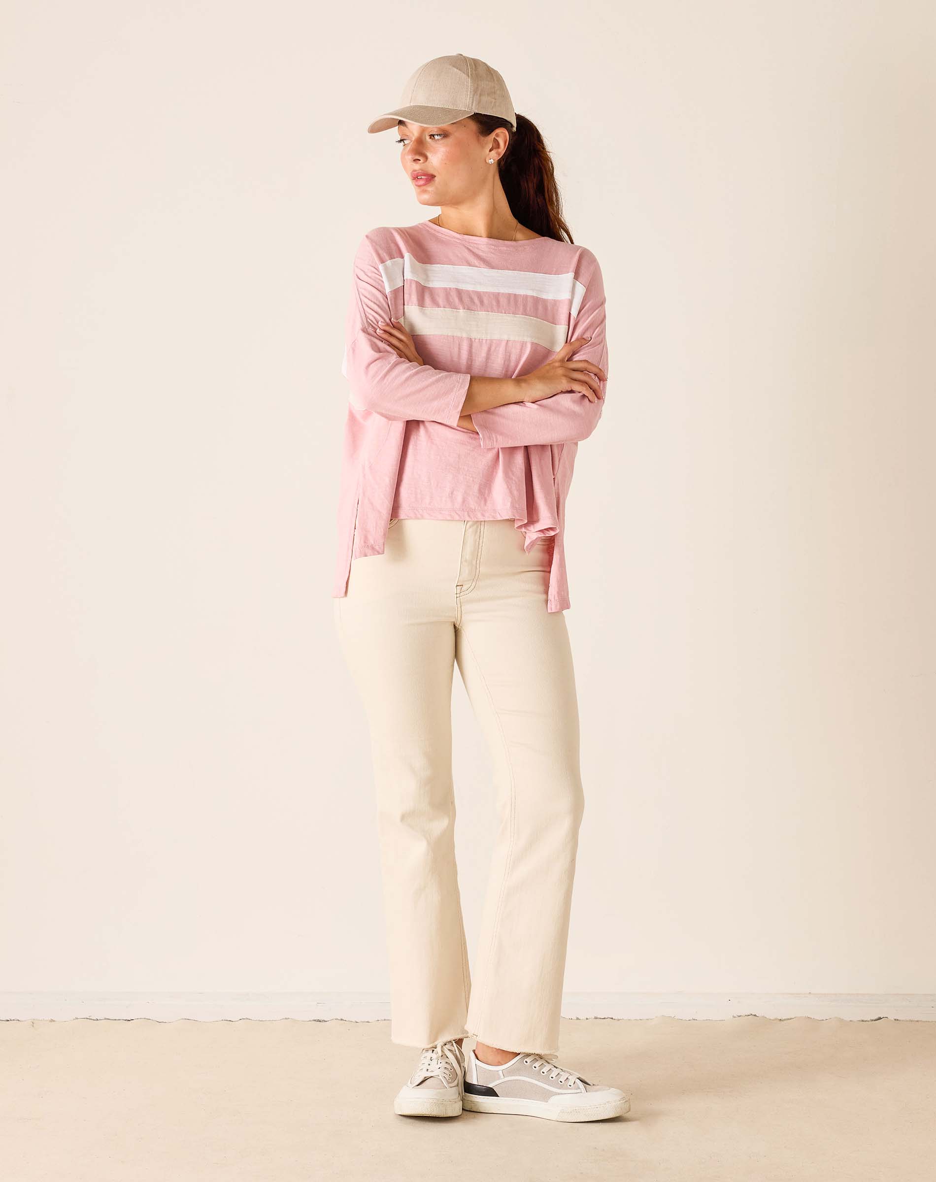 Woman wearing a pink striped sweater, beige pants, and a cap against a plain background