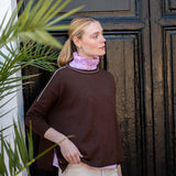 Model wearing a coffee-colored knit crewneck sweater with contrast trim over a lilac ruffled shirt and cream pants.