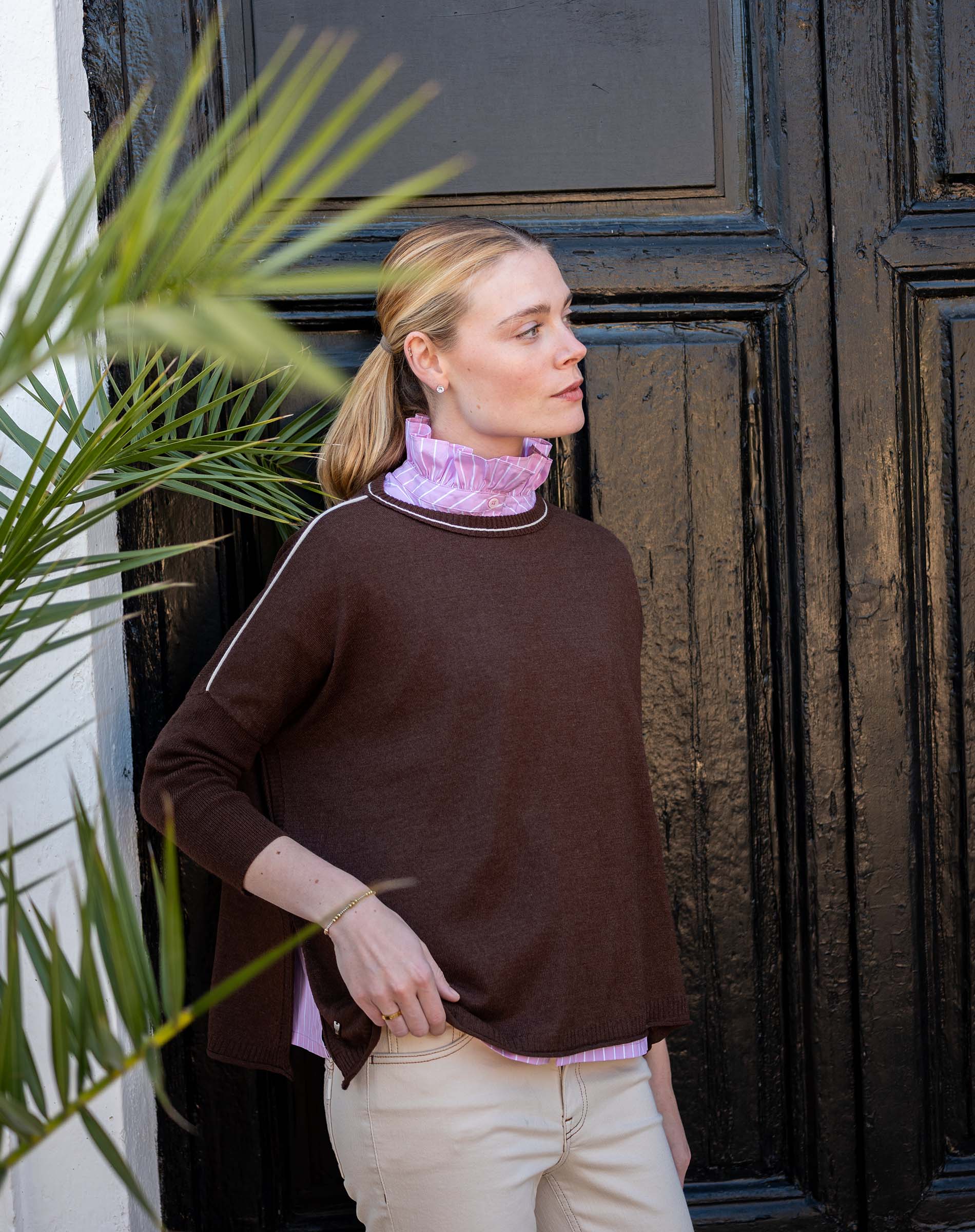 Model wearing a coffee-colored knit crewneck sweater with contrast trim over a lilac ruffled shirt and cream pants.