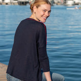 Woman in navy and red knit crewneck top with 3/4 sleeves and blue jeans sitting on a dock near water