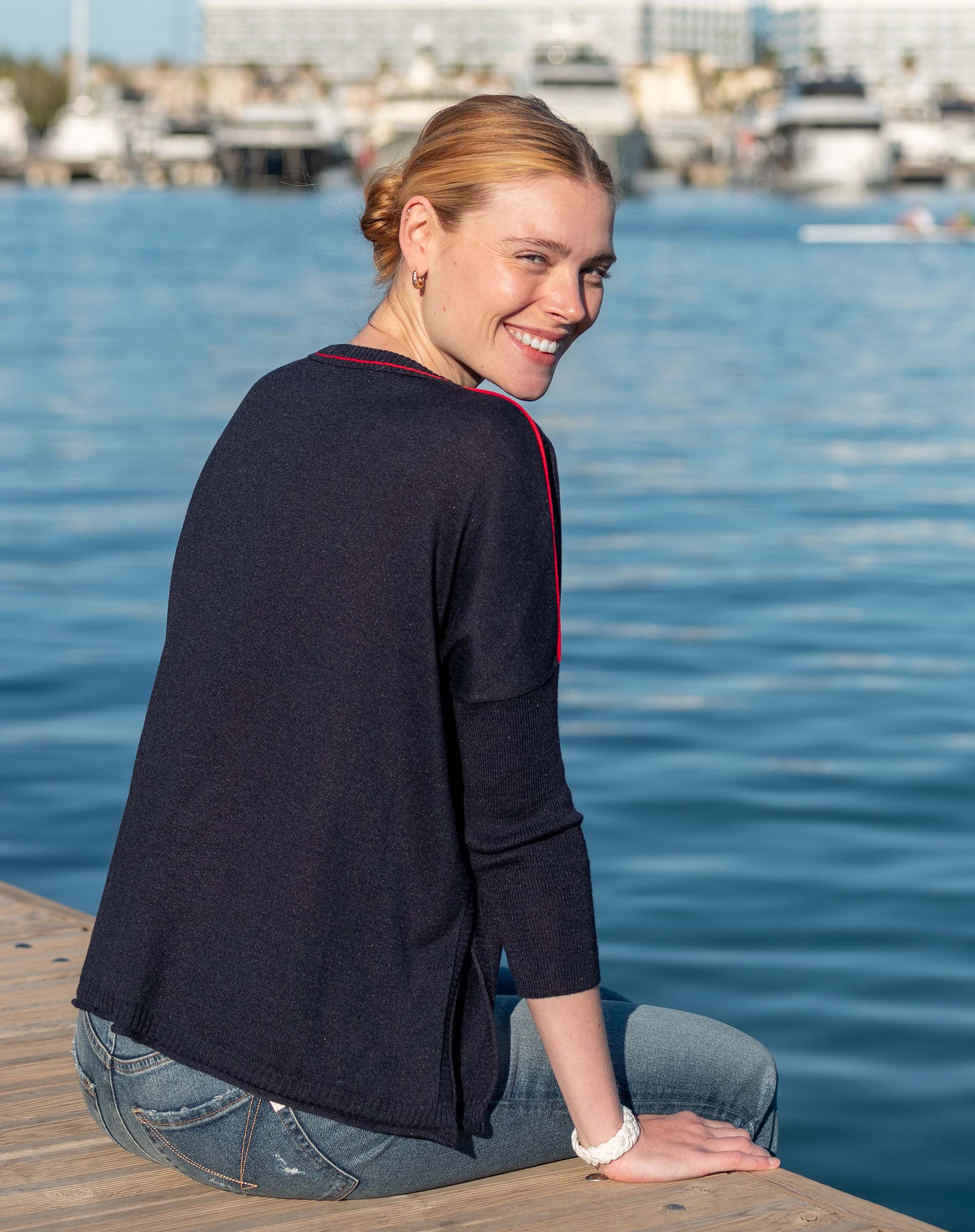 Woman in navy and red knit crewneck top with 3/4 sleeves and blue jeans sitting on a dock near water