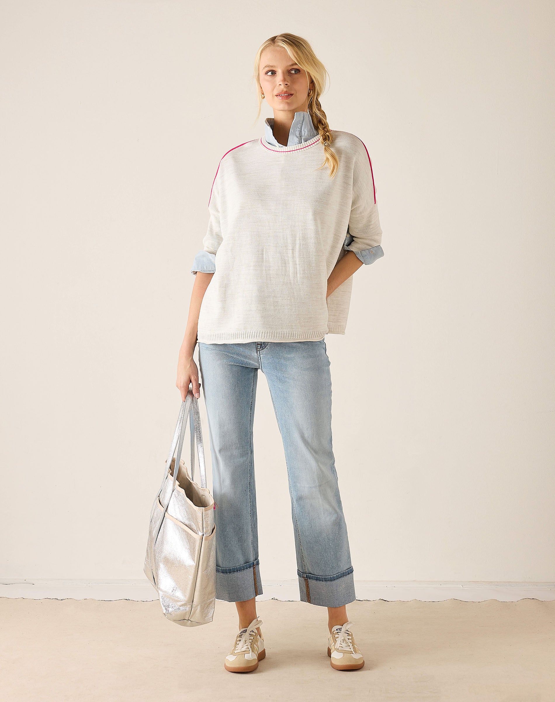 Woman wearing a light-colored sweater with rolled-up sleeves and light blue jeans, holding a silver tote bag.