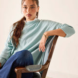 Woman wearing a light blue sweater and blue jeans sitting on a wooden chair.