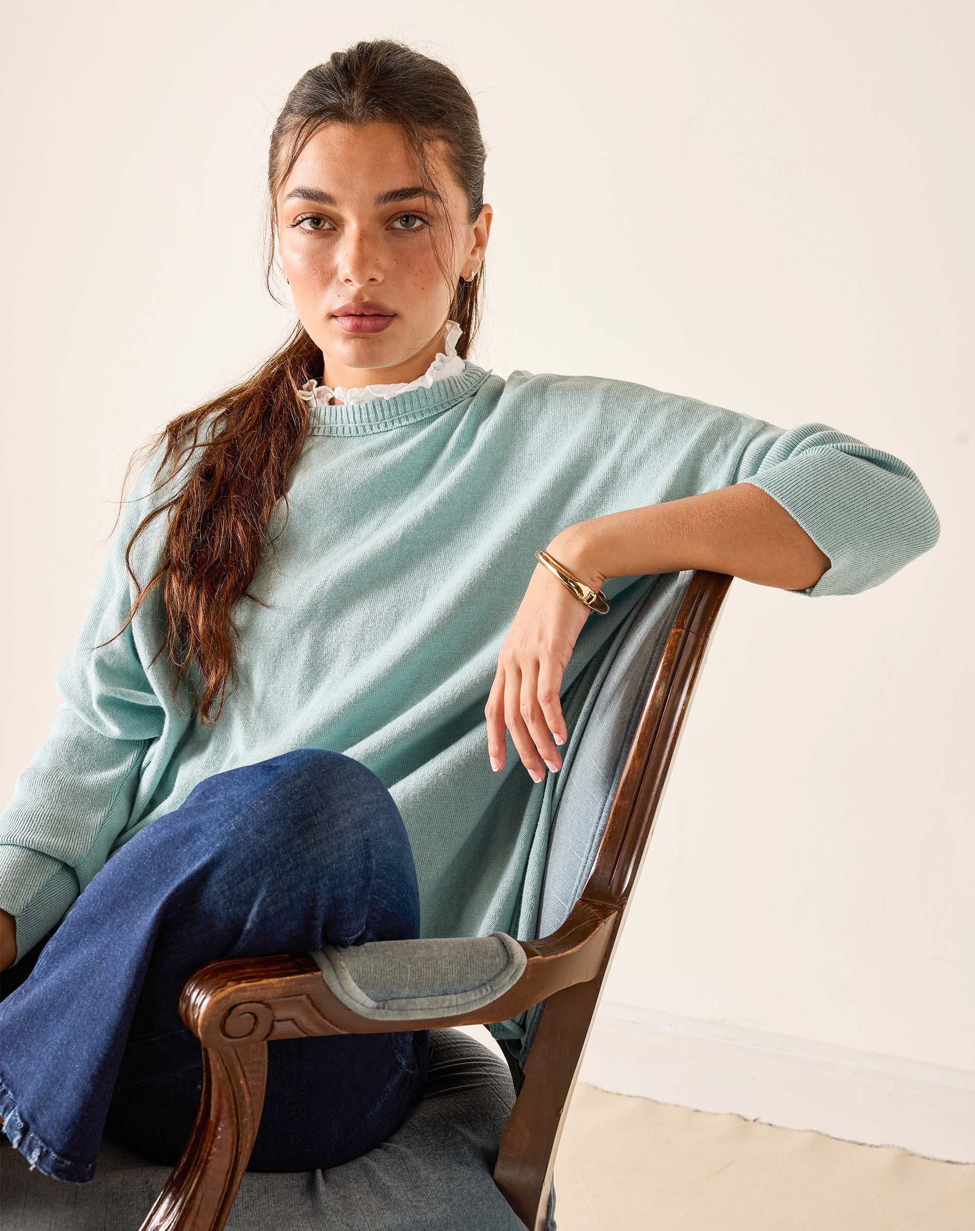Woman wearing a light blue sweater and blue jeans sitting on a wooden chair.
