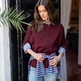 Burgundy knit ruffle sweater with striped scarf tied at the waist worn over a light blue shirt and jeans.