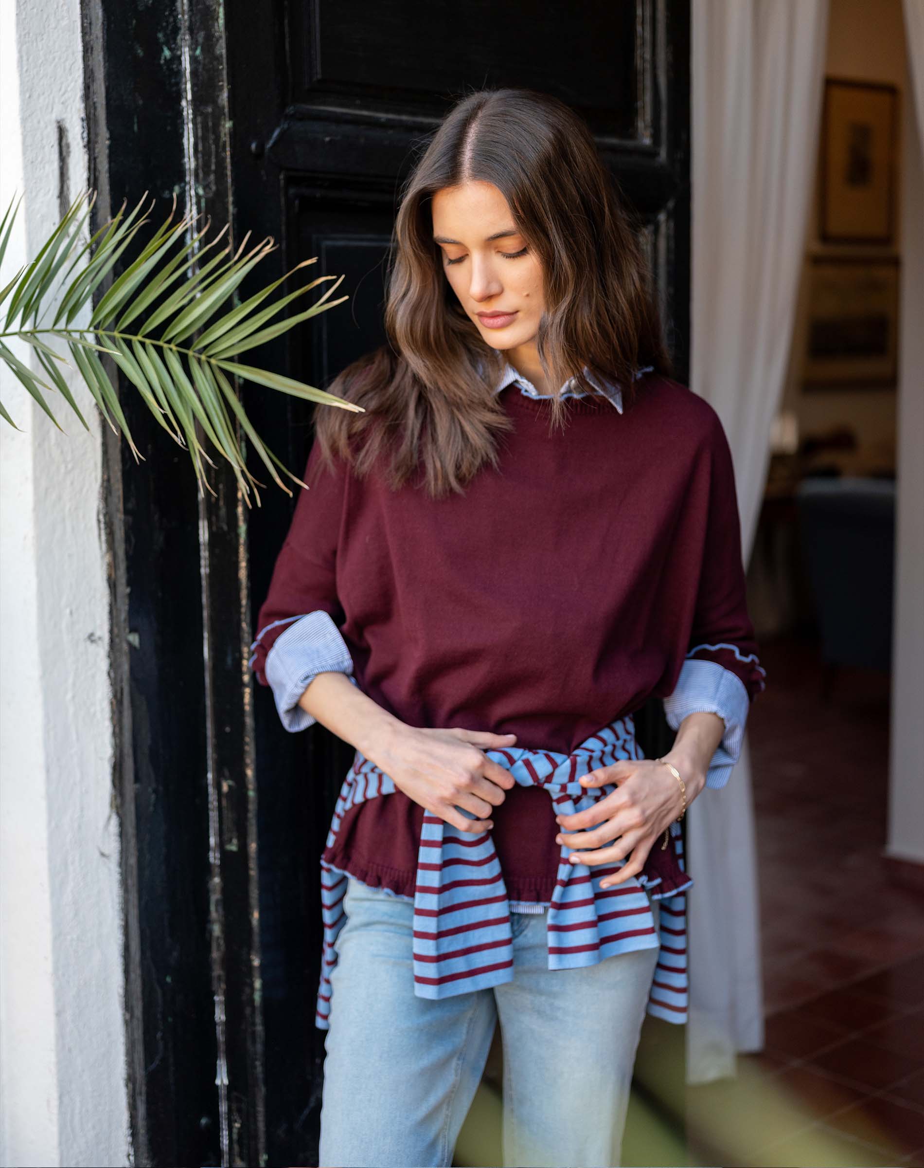Burgundy knit ruffle sweater with striped scarf tied at the waist worn over a light blue shirt and jeans.