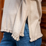 Natural cotton ruffled sweater with a side slit and fringed hem, worn over blue denim jeans.