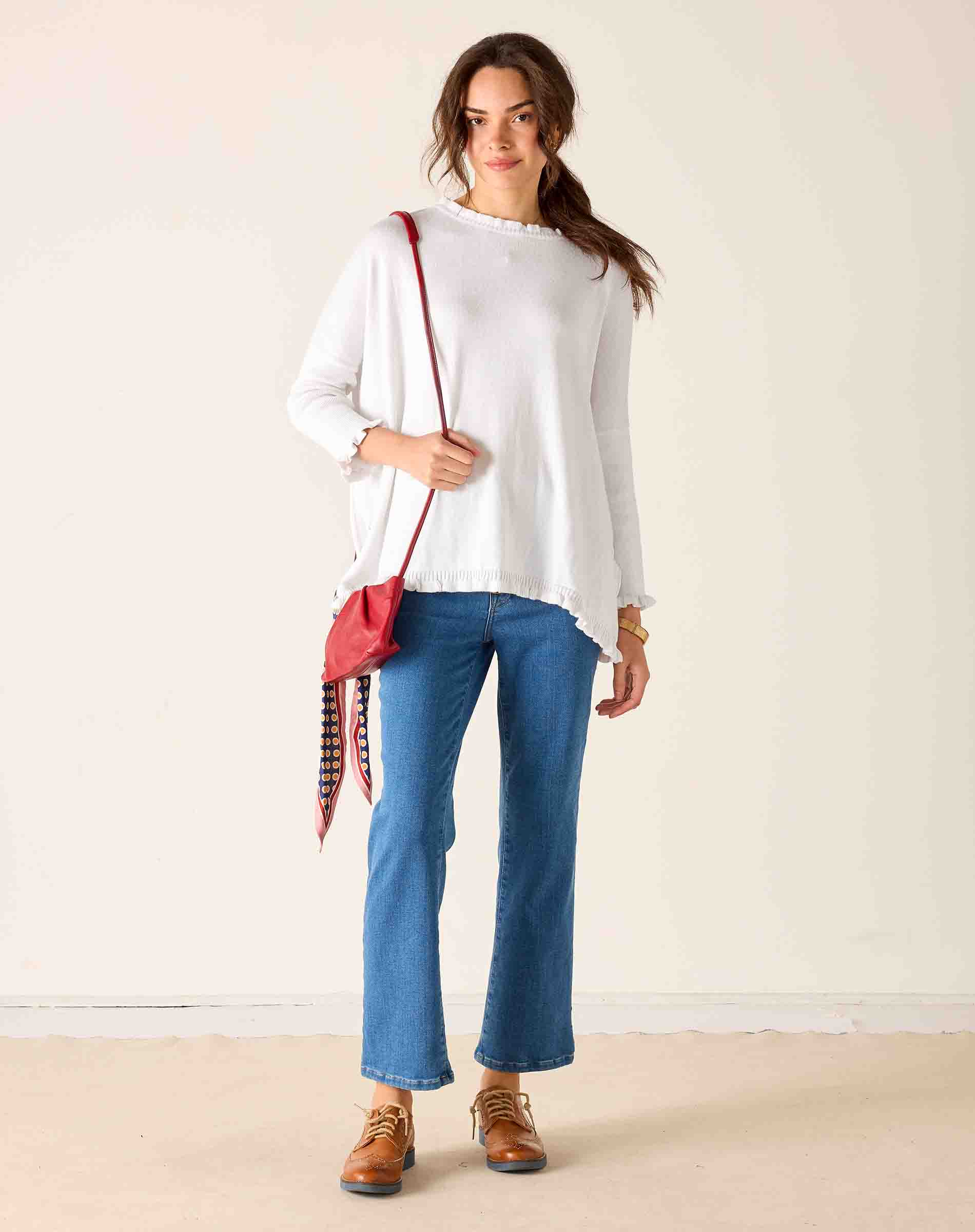 Woman wearing a white top, blue jeans, and brown shoes with a red bag against a beige background