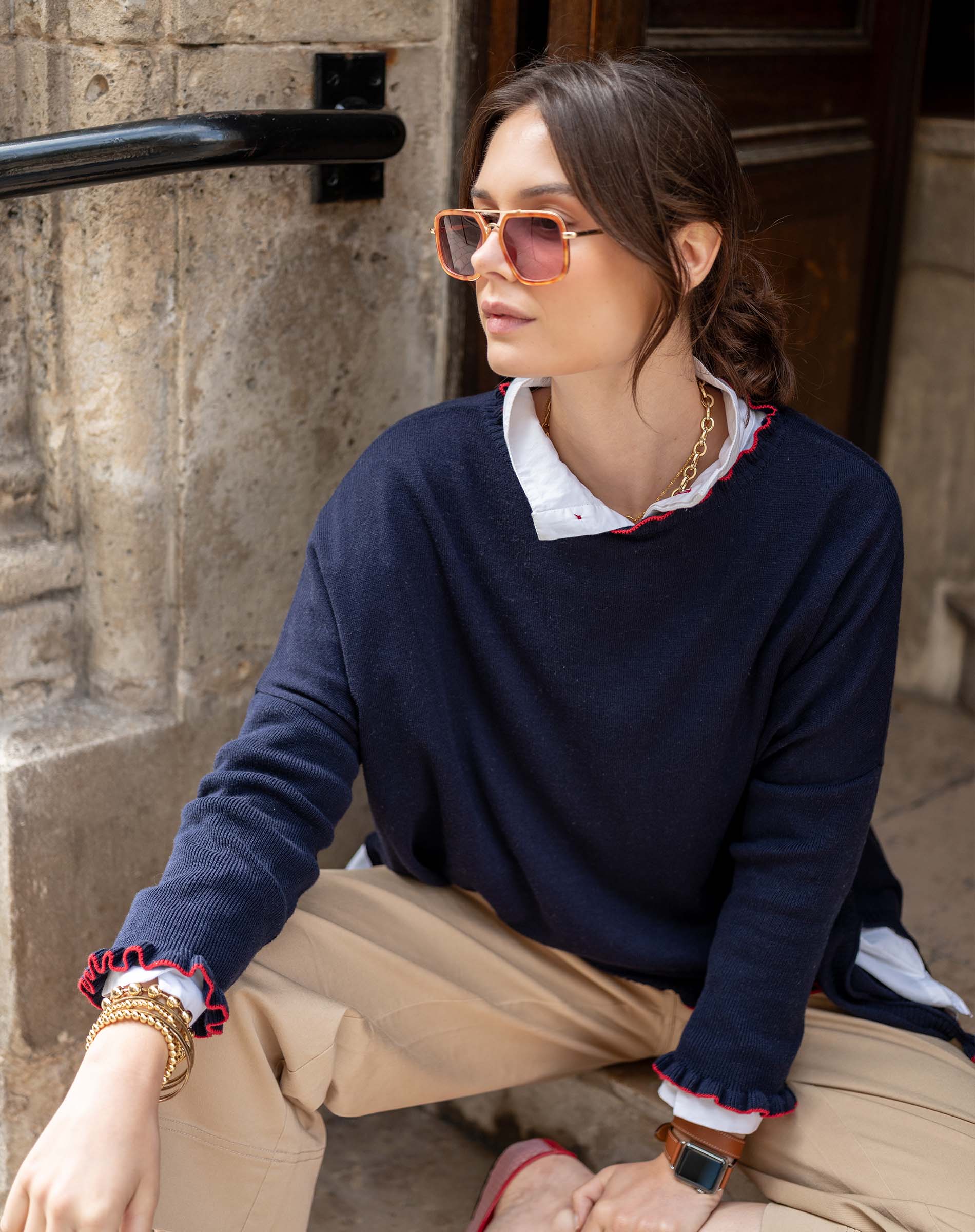 Woman wearing a navy sweater with ruffled cuffs and hem, sitting against a stone wall.
