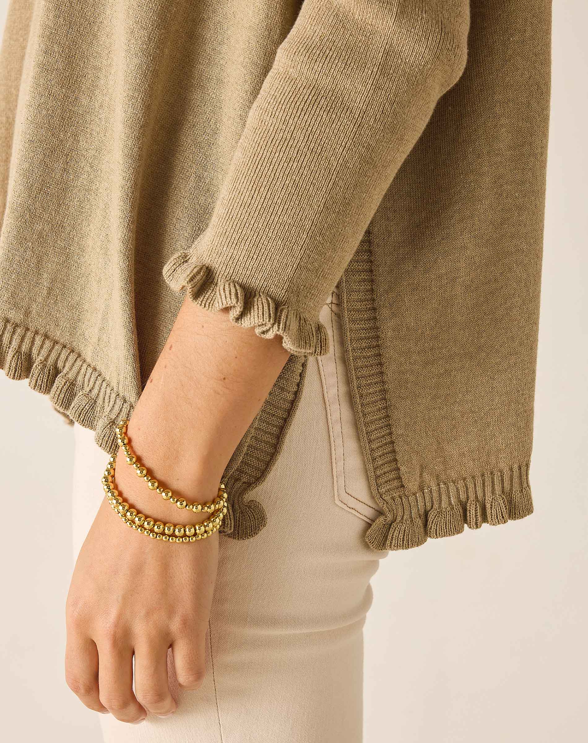 Close-up of a person wearing a beige sweater with ruffled cuffs and a bracelet on a neutral background