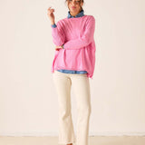 Woman wearing a pink sweater over a blue shirt with white pants on a plain background