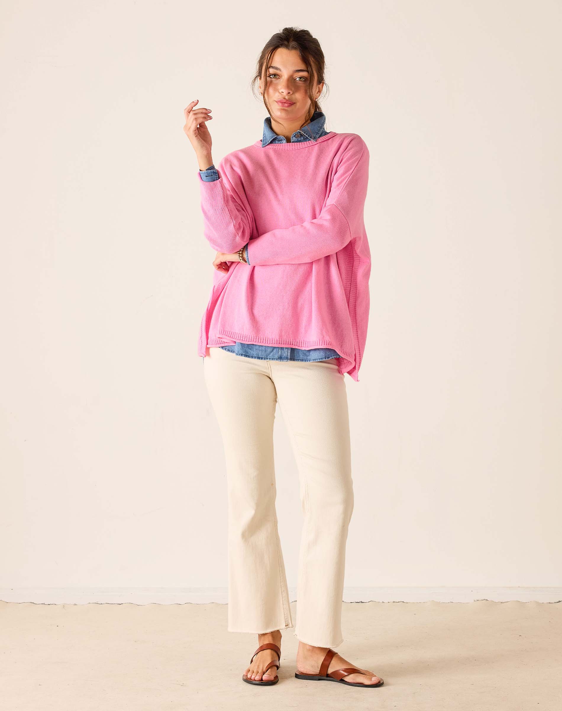 Woman wearing a pink sweater over a blue shirt with white pants on a plain background