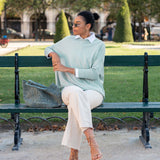 Woman sitting on a bench wearing a light green sweater and white pants in an outdoor setting.
