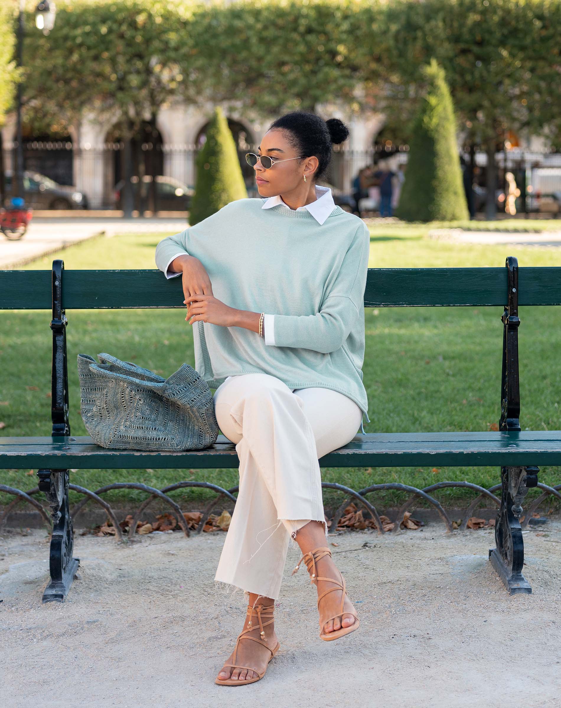 Woman sitting on a bench wearing a light green sweater and white pants in an outdoor setting.