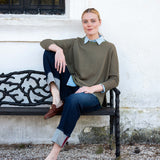Model wearing an olive green crewneck knit top with a collared shirt underneath and dark denim jeans.