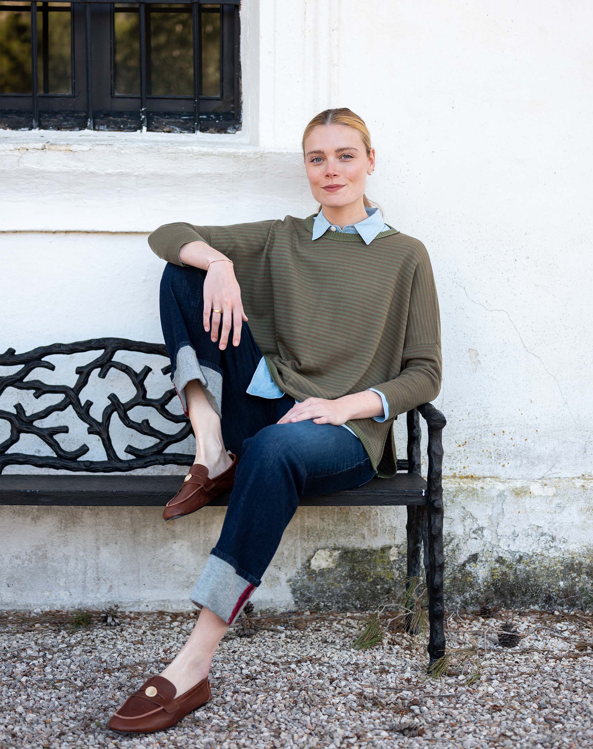 Model wearing an olive green crewneck knit top with a collared shirt underneath and dark denim jeans.