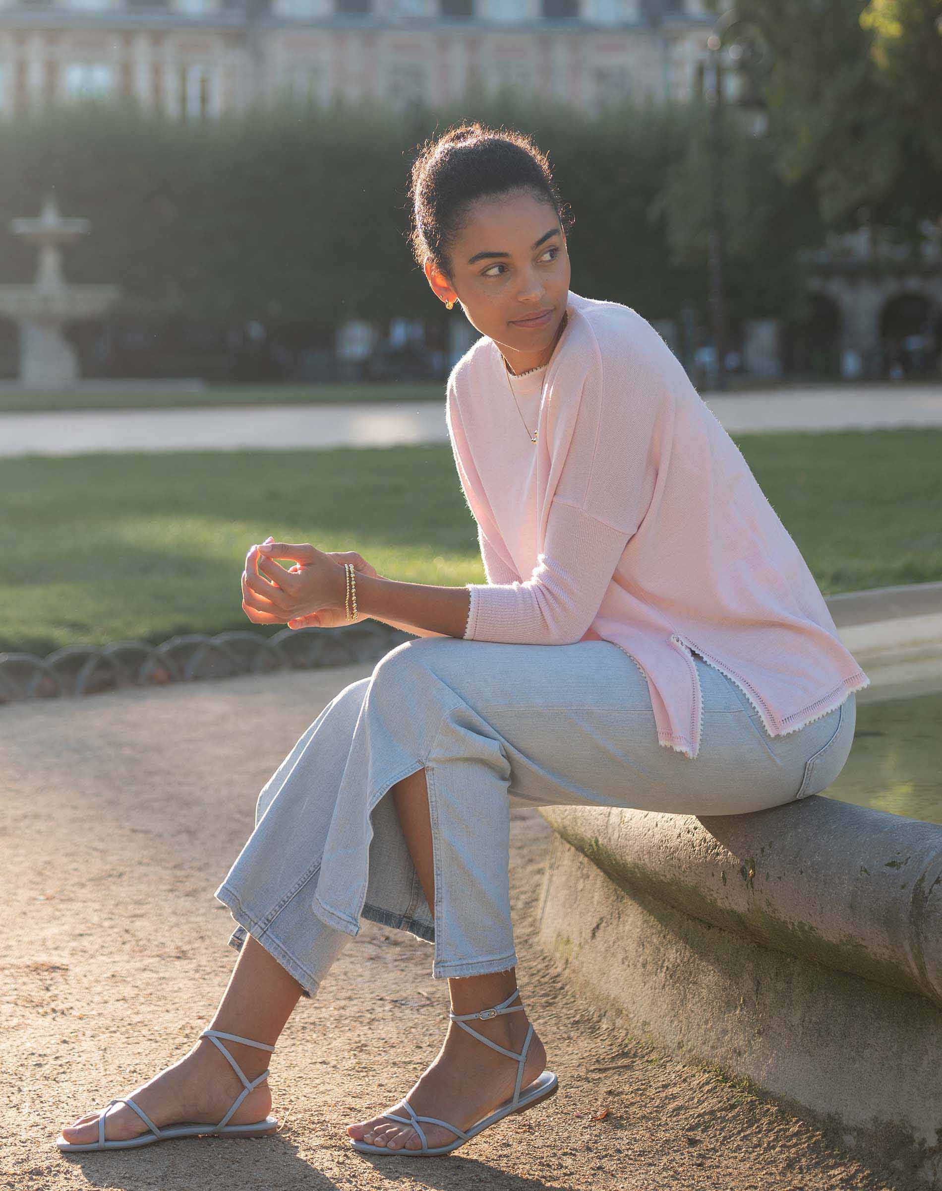 Woman sitting on a stone bench in a park wearing a pink cardigan and light blue pants.