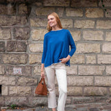 Woman in a blue sweater and white pants standing against a stone wall.