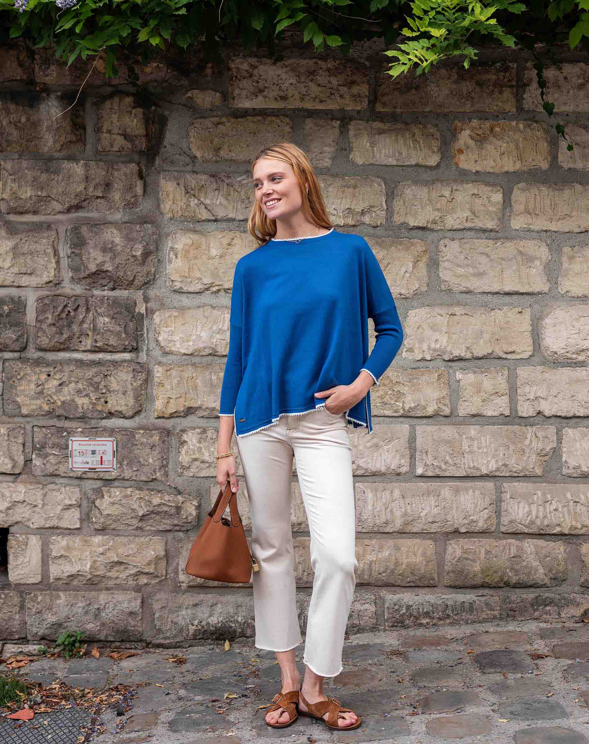 Woman in a blue sweater and white pants standing against a stone wall.