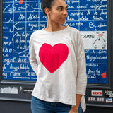 Woman wearing a white sweatshirt with a red heart design in front of a blue wall with white text.