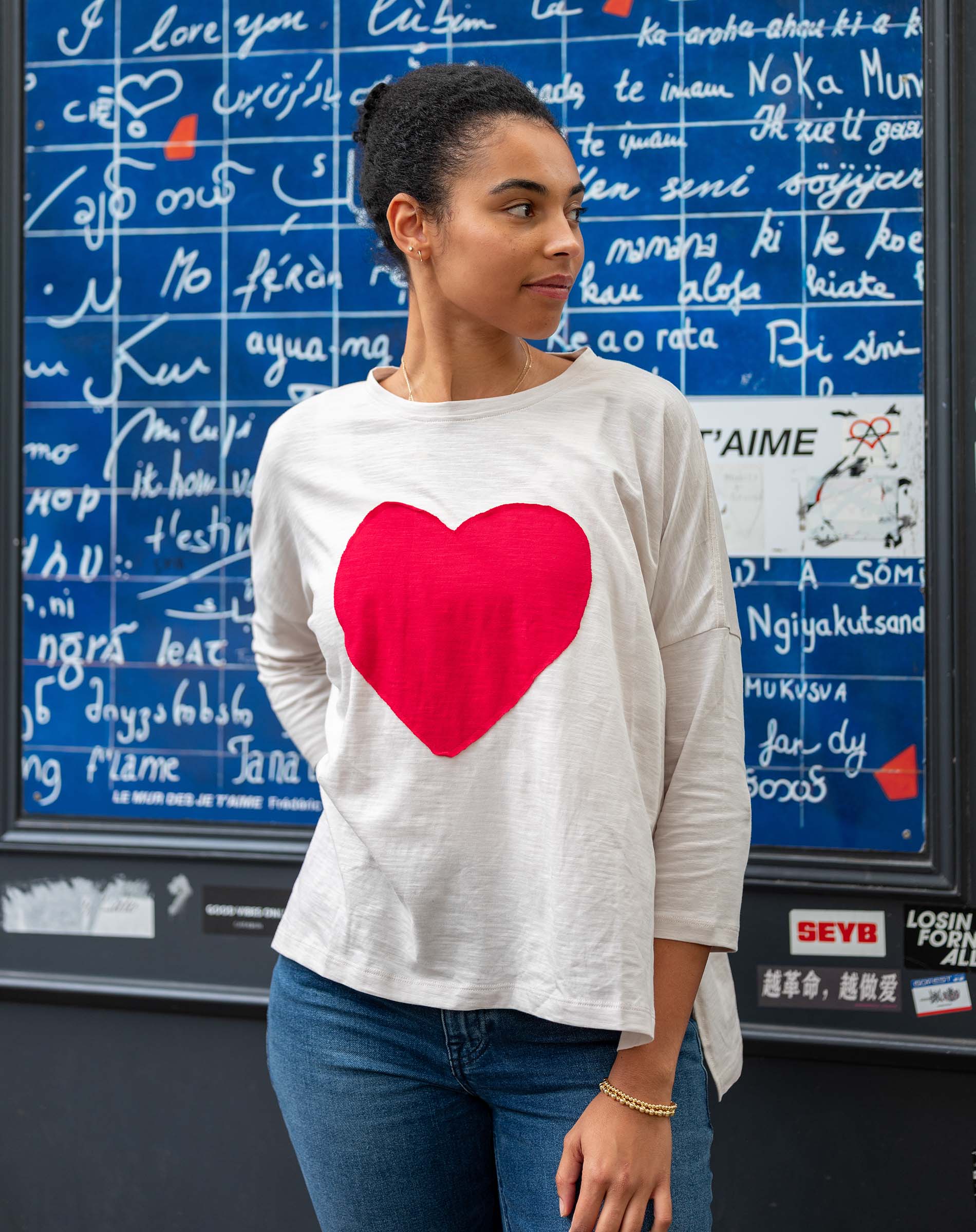 Woman wearing a white sweatshirt with a red heart design in front of a blue wall with white text.
