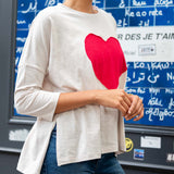 Person wearing a white long-sleeve shirt with a red heart design in front of a blue board with writing.