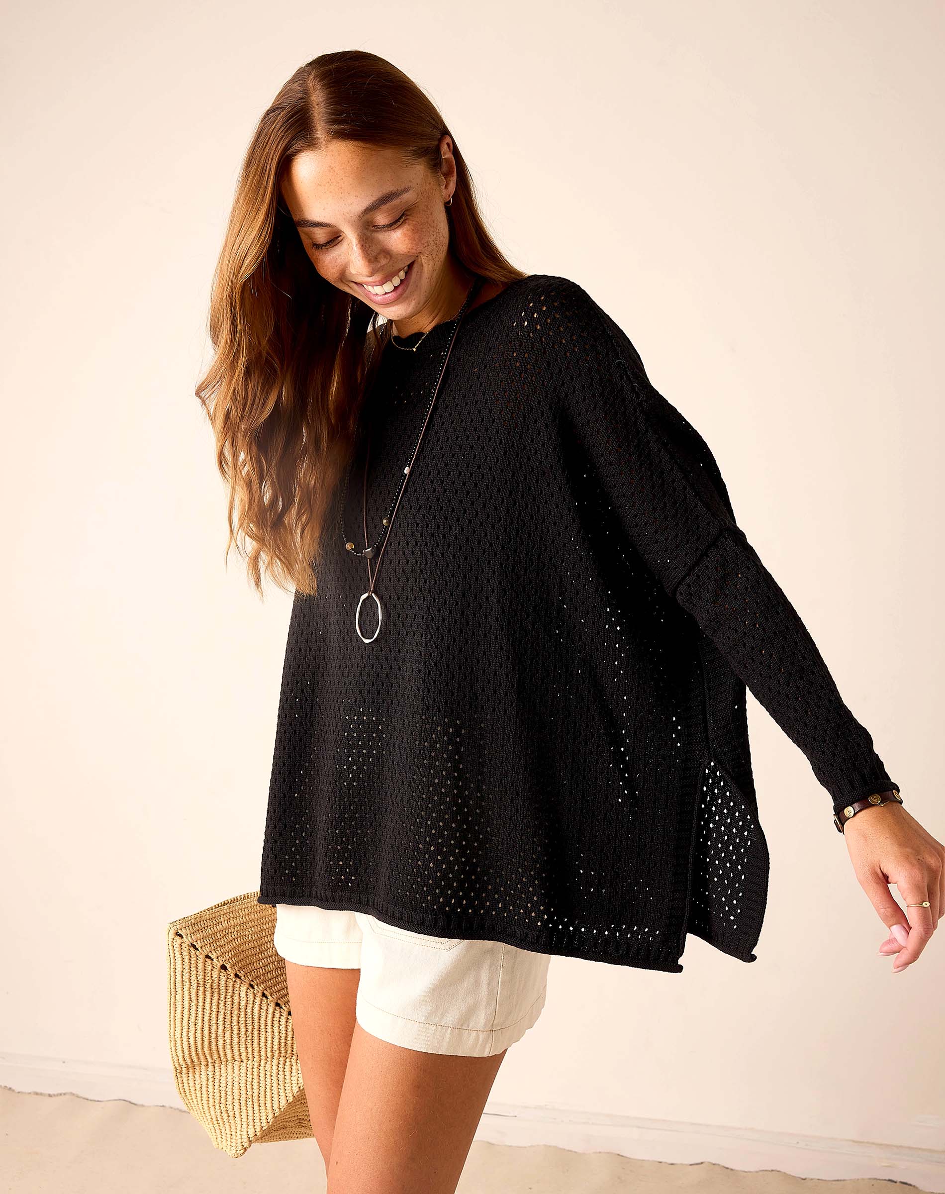 Woman wearing a black perforated sweater and white shorts, holding a woven bag.