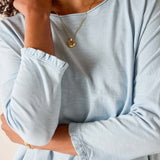 Person wearing a light blue long-sleeve shirt with rolled-up sleeves, gold necklace, and bracelet.