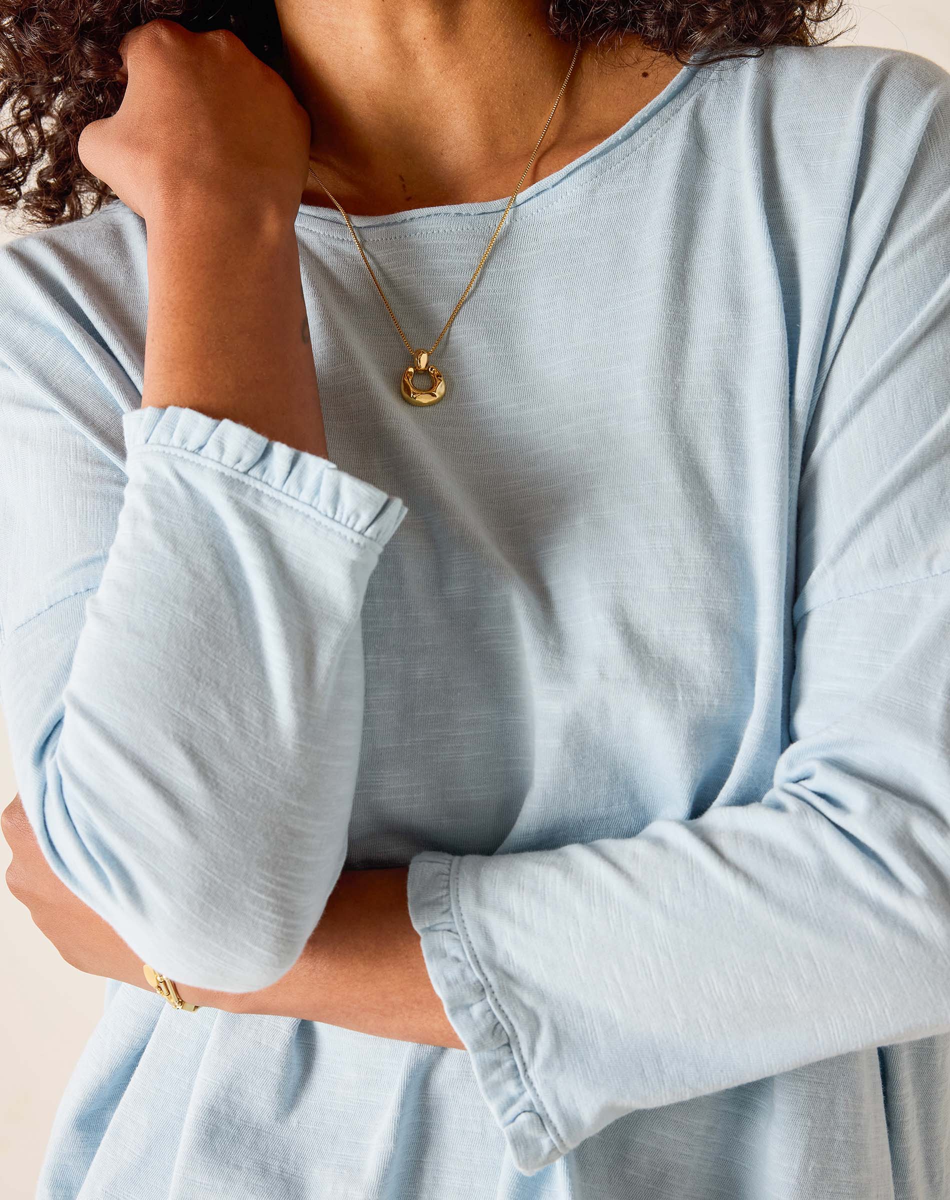 Person wearing a light blue long-sleeve shirt with rolled-up sleeves, gold necklace, and bracelet.