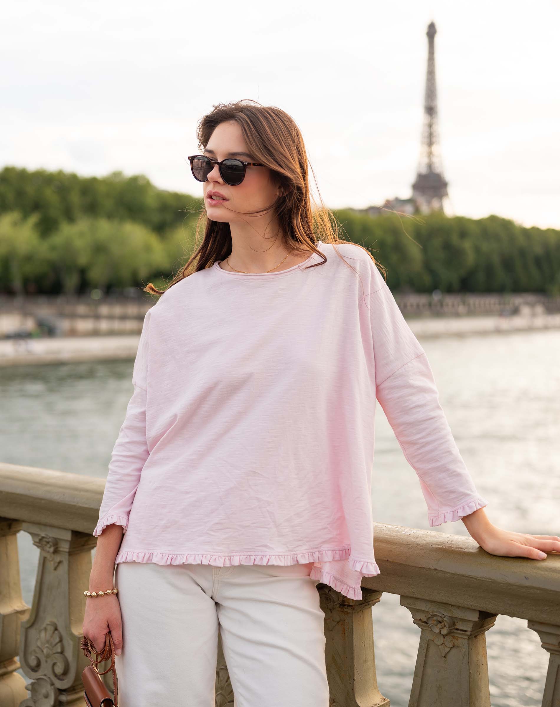 Woman in a pink top and white pants standing by a waterfront with the Eiffel Tower in the background.