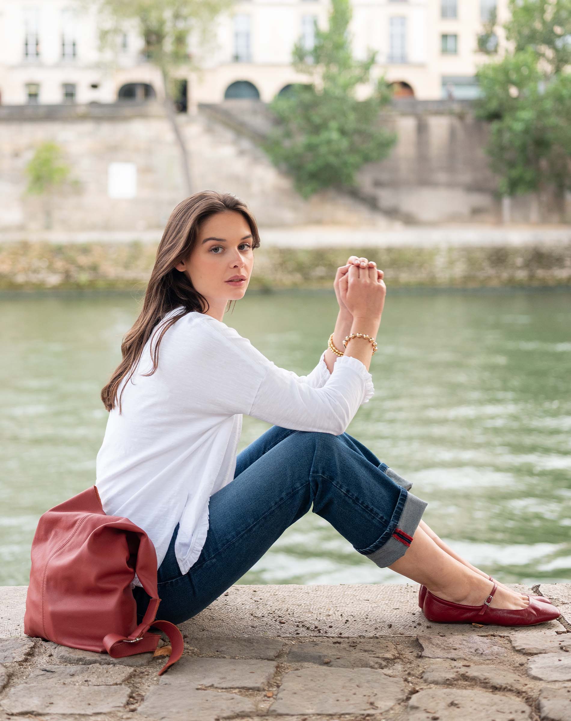 Woman sitting by a river with a red bag, wearing a white shirt and blue jeans.
