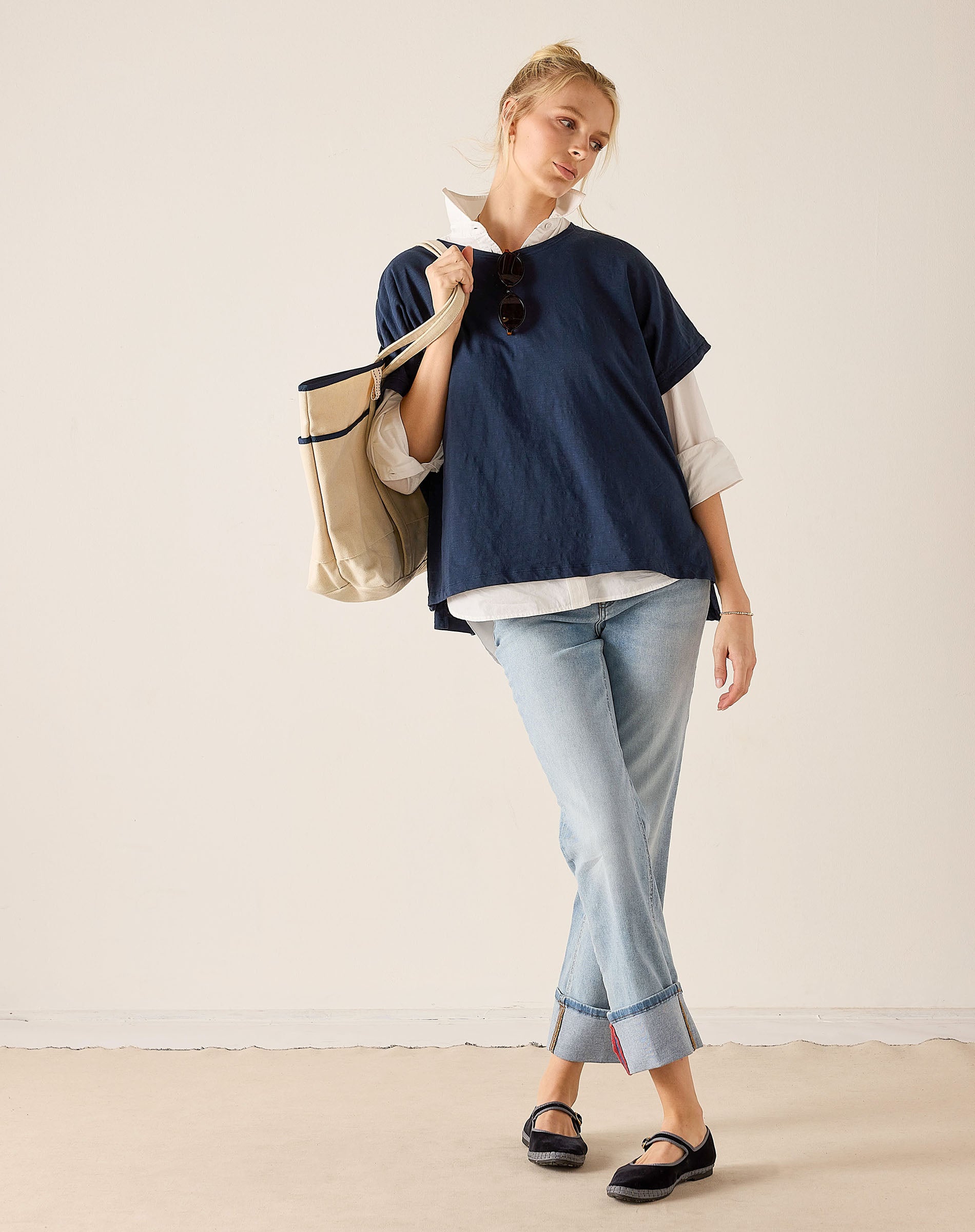 Woman wearing a navy blue top and light blue jeans, holding a beige tote bag against a plain background.