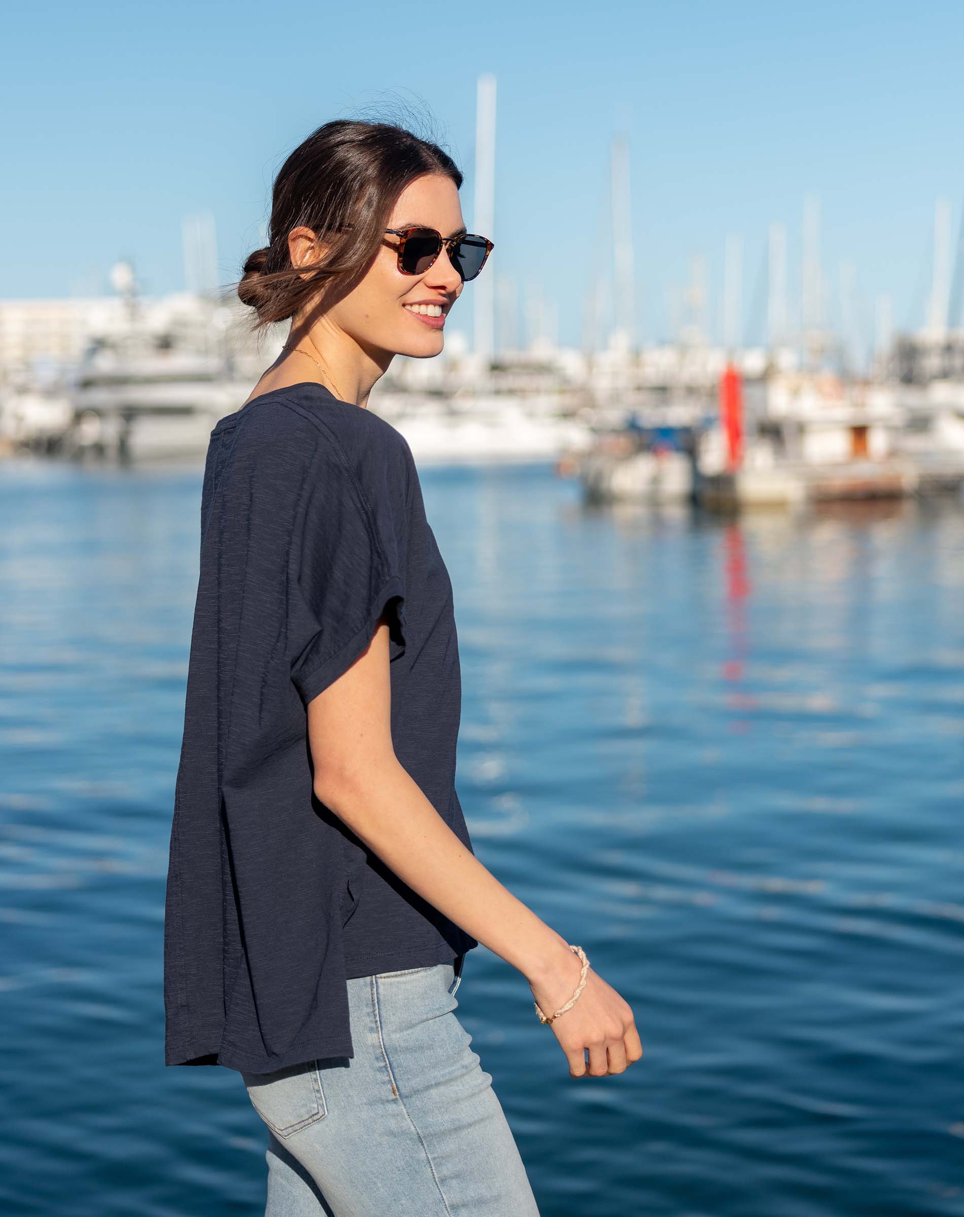 Woman in a dark navy linen shirt & light wash jeans by MERSEA, overlooking a harbor. Coastal travel vibes.