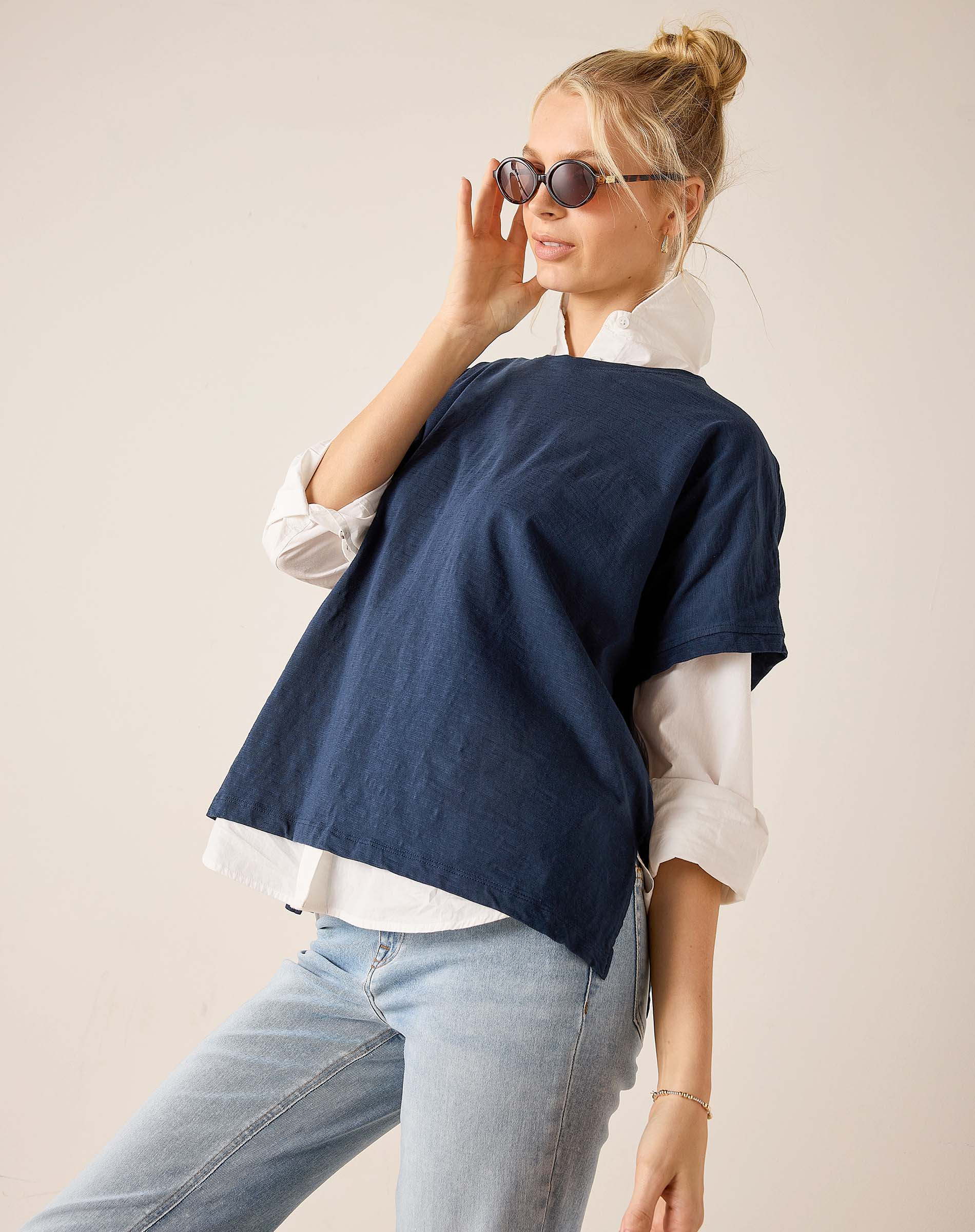 Woman wearing a navy blue poncho over a white shirt and light blue jeans, holding sunglasses.