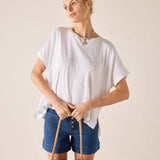 Woman wearing a white oversized shirt and blue shorts, holding a woven bag against a beige background