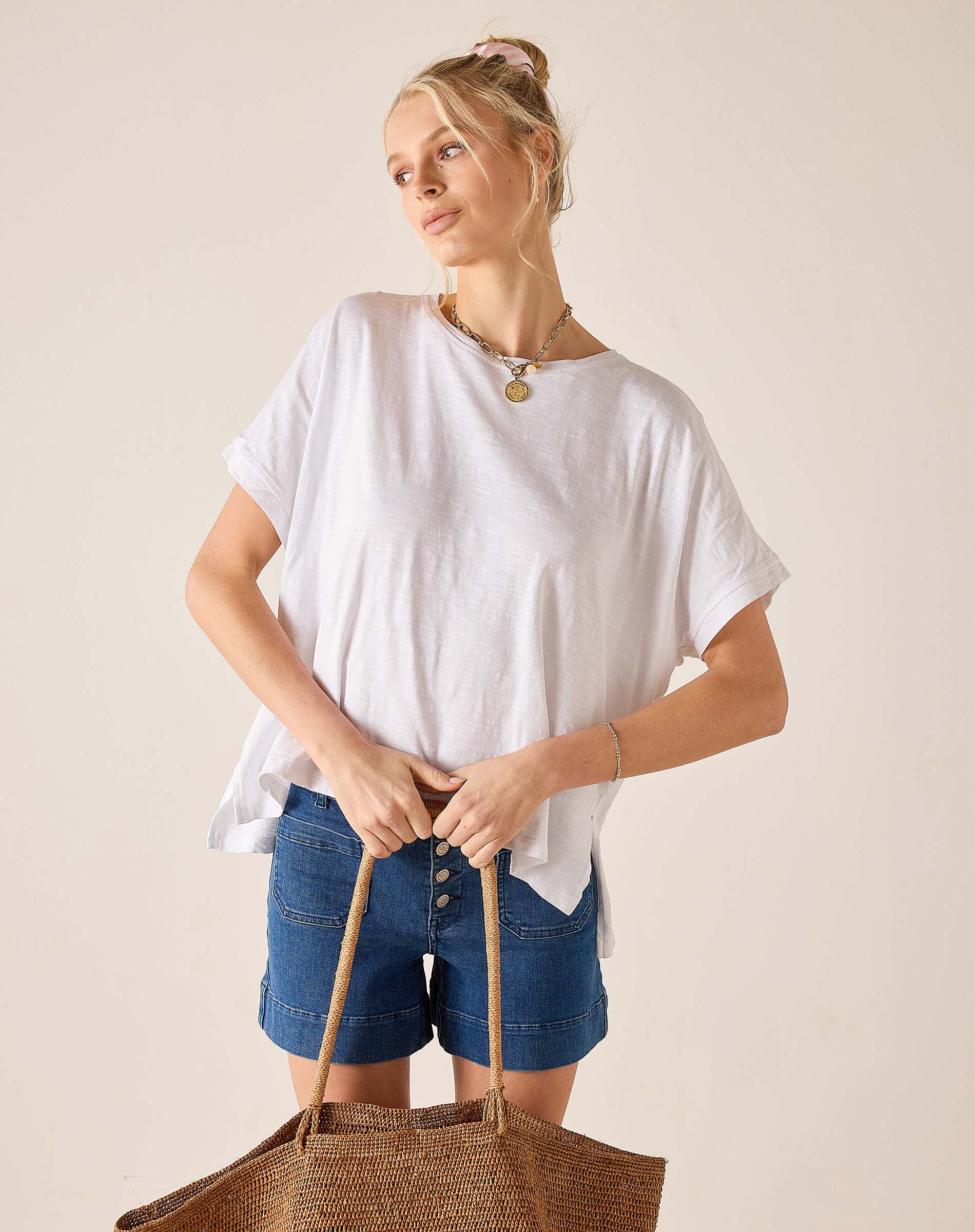 Woman wearing a white oversized shirt and blue shorts, holding a woven bag against a beige background