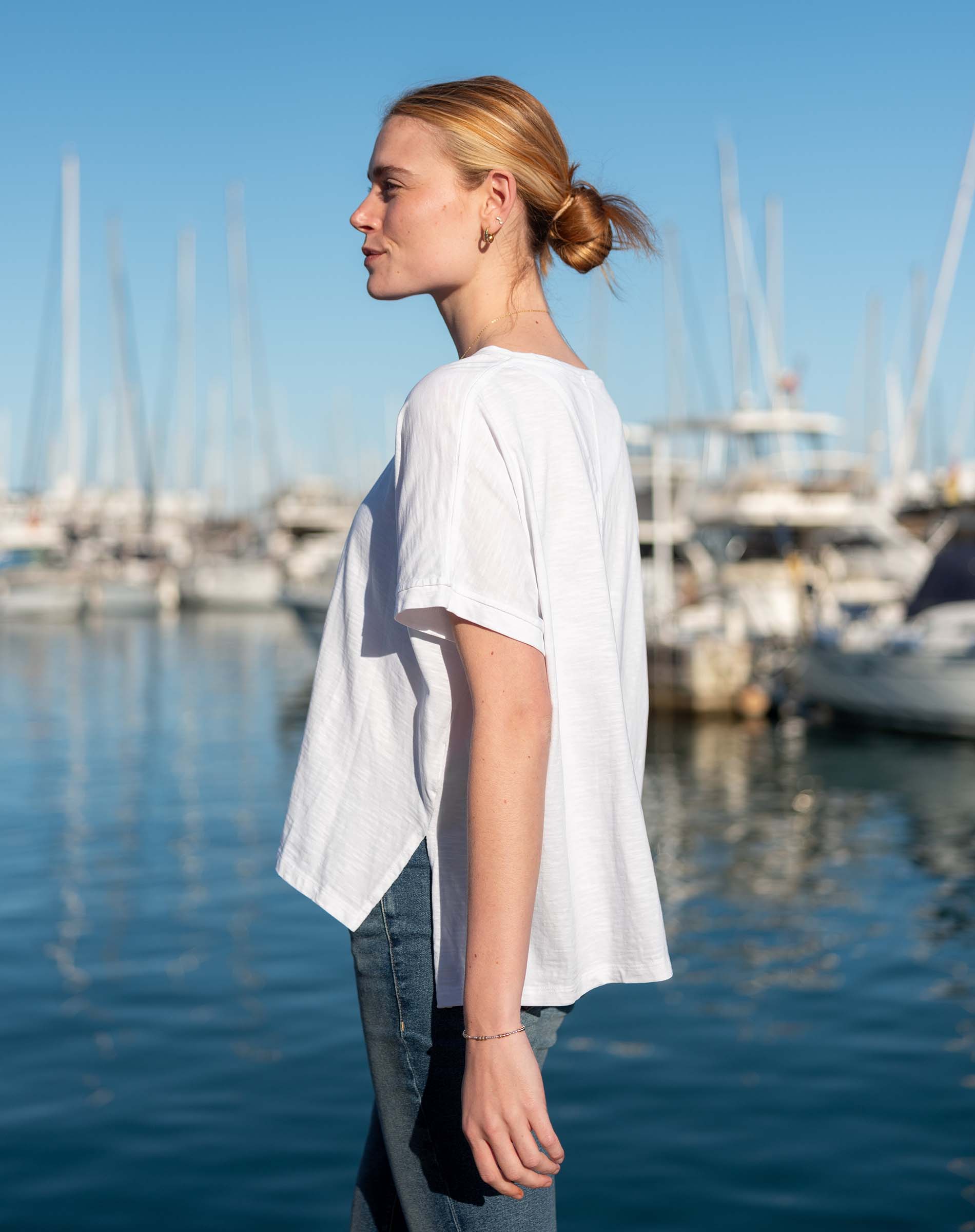 Woman in white short-sleeve knit tee and dark denim jeans standing in front of a harbor.