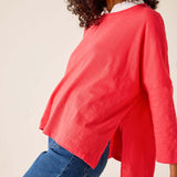 Person wearing a red sweater over a white shirt with blue jeans on a plain background