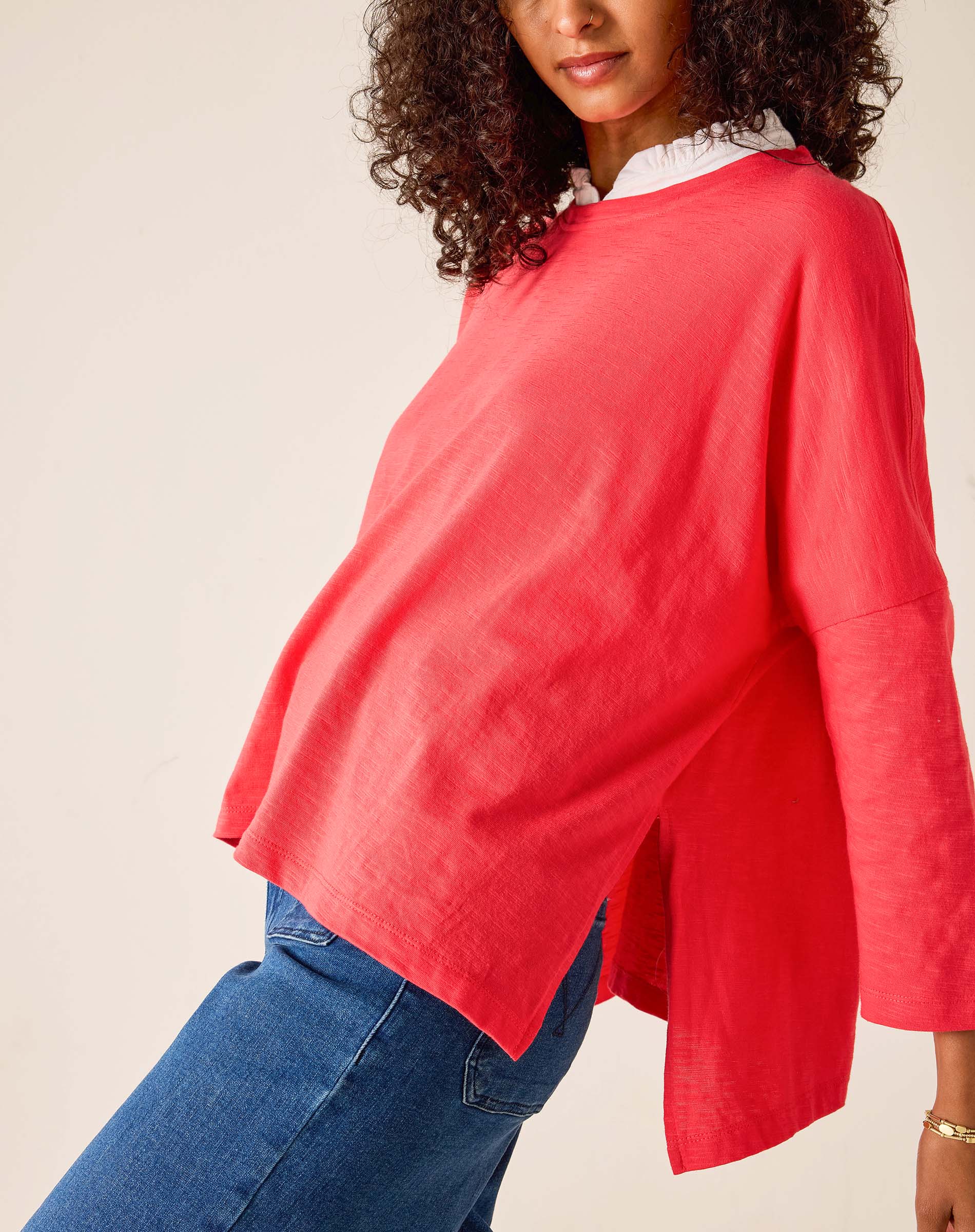 Person wearing a red sweater over a white shirt with blue jeans on a plain background