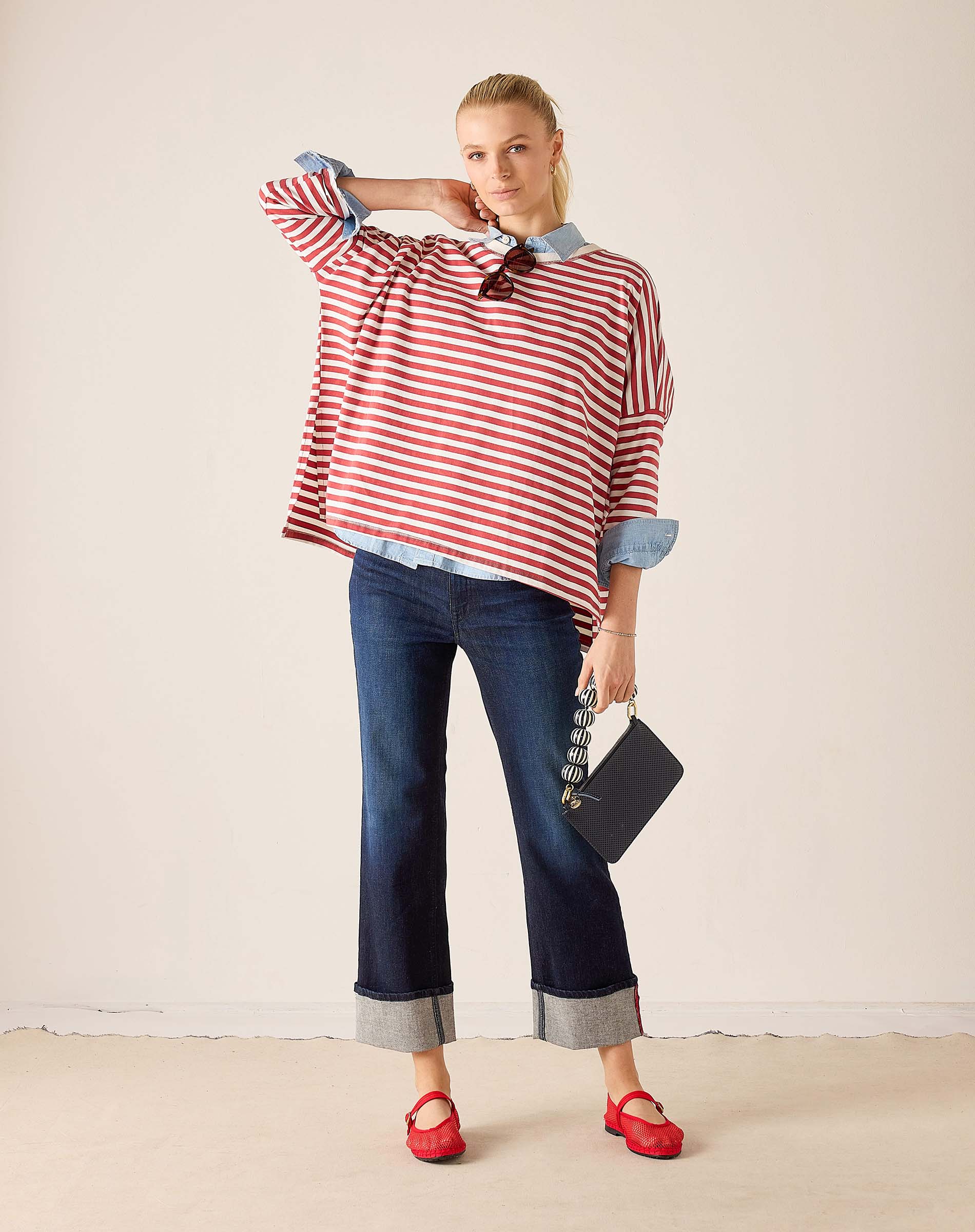 Woman wearing a red and white striped shirt with rolled-up sleeves, blue jeans, and red shoes, holding a black clutch.