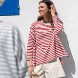 Woman wearing a red and white striped shirt and beige shorts against a white wall with blue shutters.