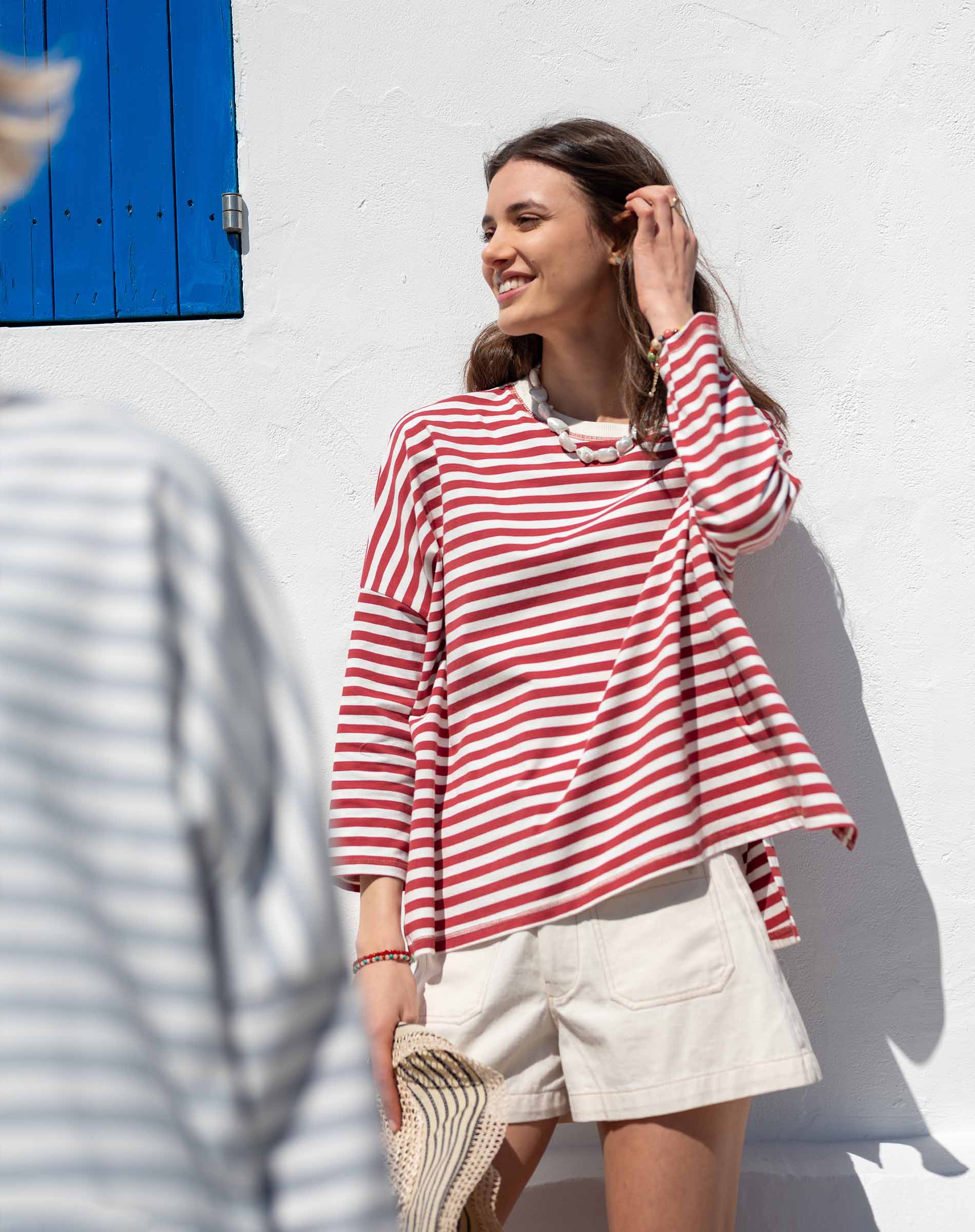 Woman wearing a red and white striped shirt and beige shorts against a white wall with blue shutters.