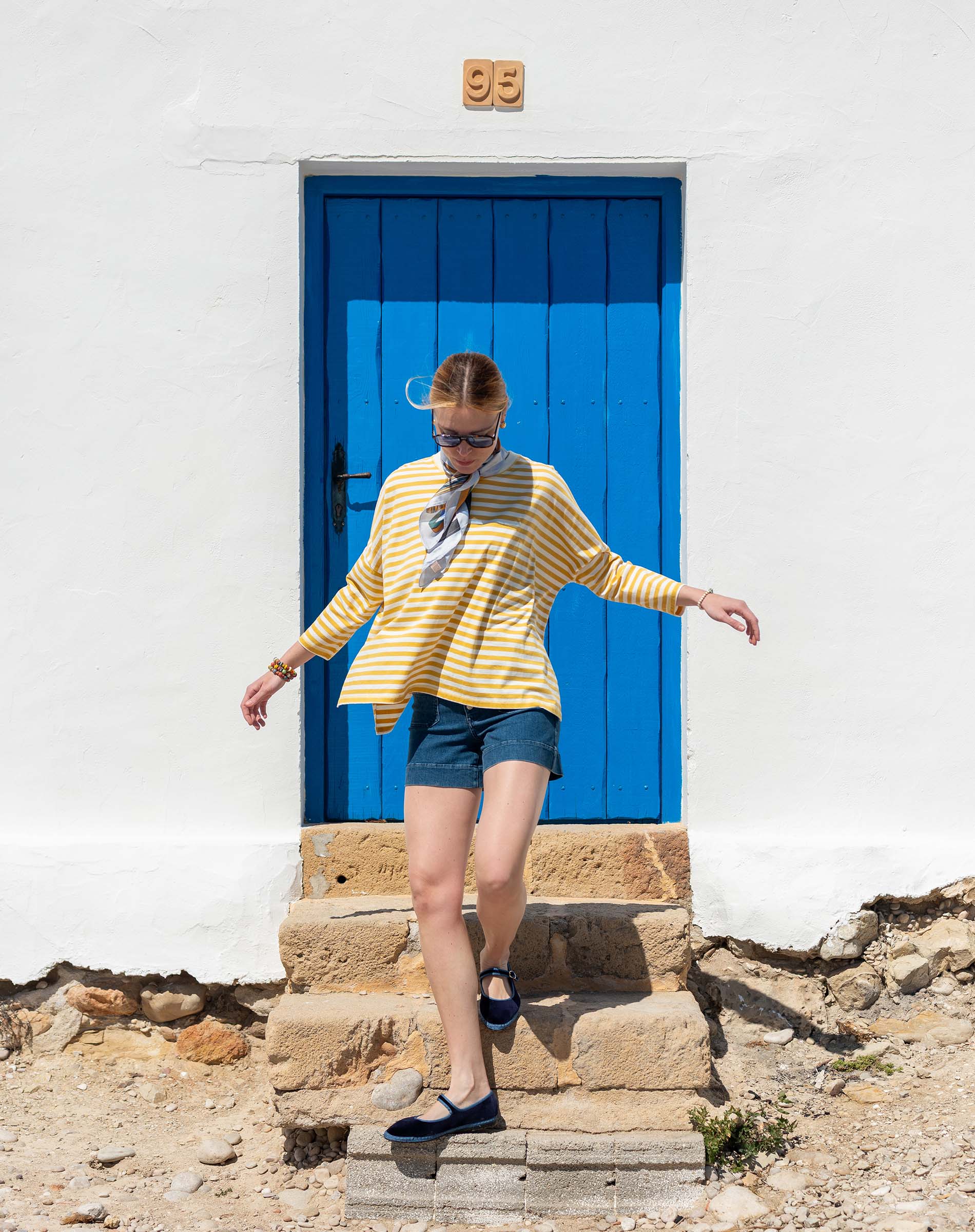 Woman in a yellow striped shirt and denim shorts standing in front of a blue door.
