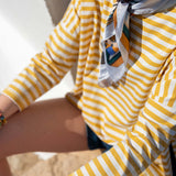 Person wearing a yellow and white striped shirt with a colorful scarf, sitting on sand.