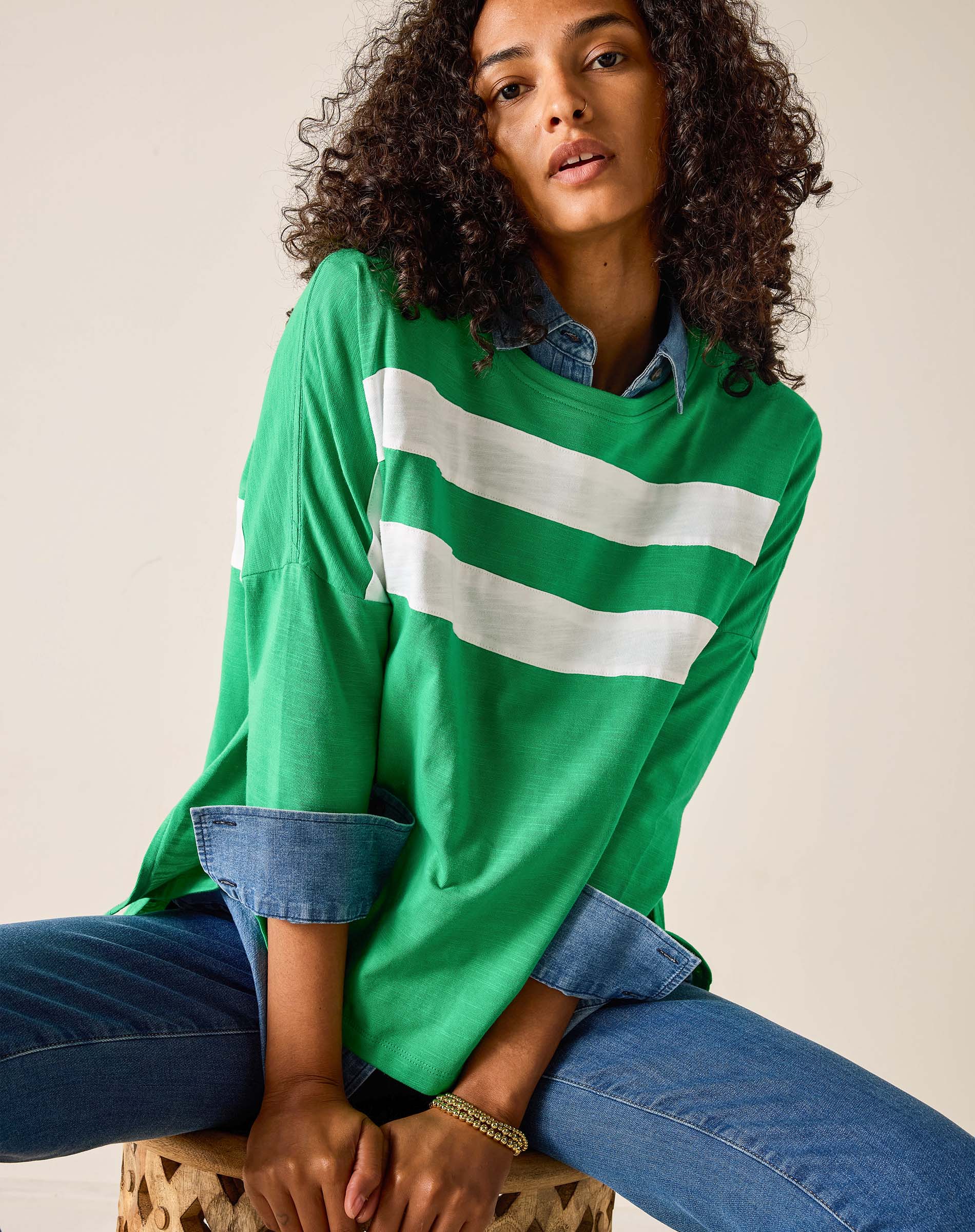Woman wearing a green oversized shirt with white stripes and blue jeans, sitting on a beige background.