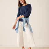 Woman wearing a navy top and white pants with a light blue shirt tied around her waist against a plain background